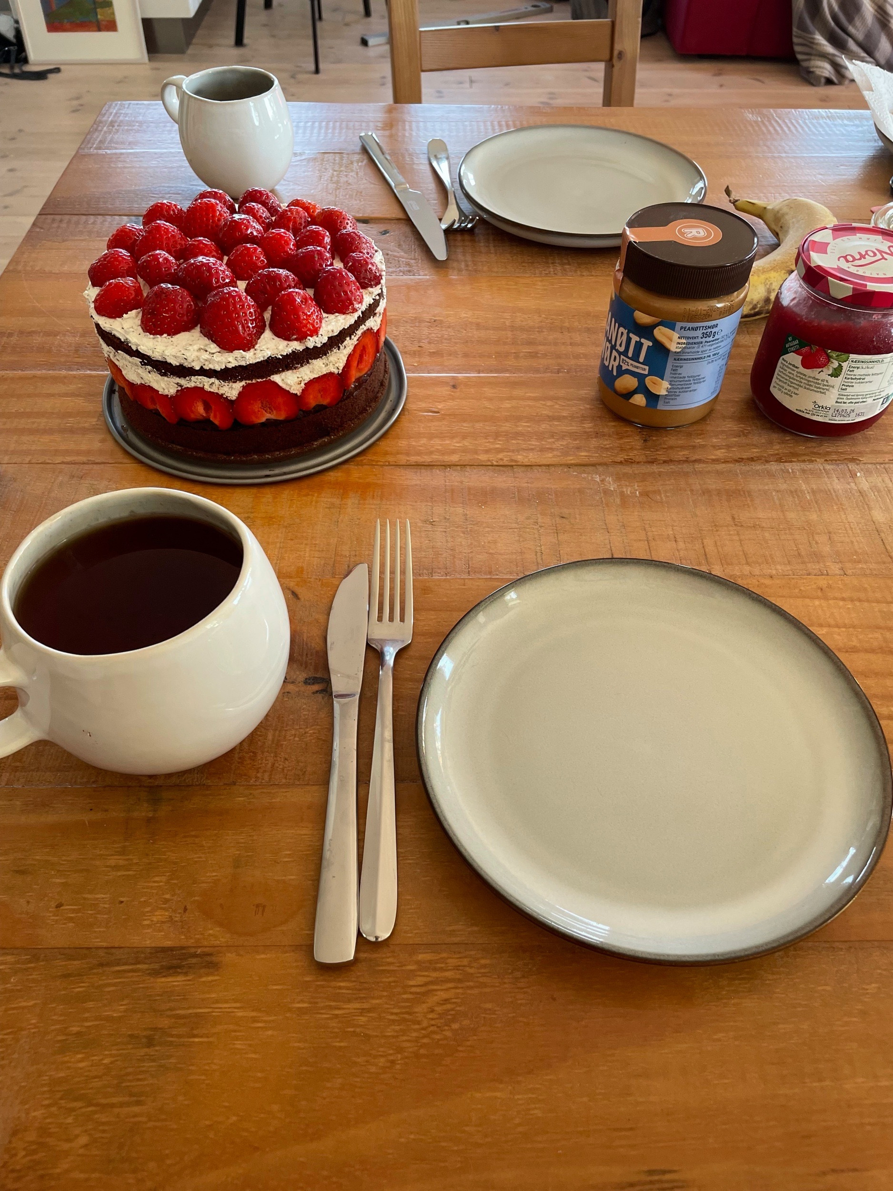 A table is set with a strawberry-topped cake, plates, utensils, a cup of tea, peanut butter, and jam.